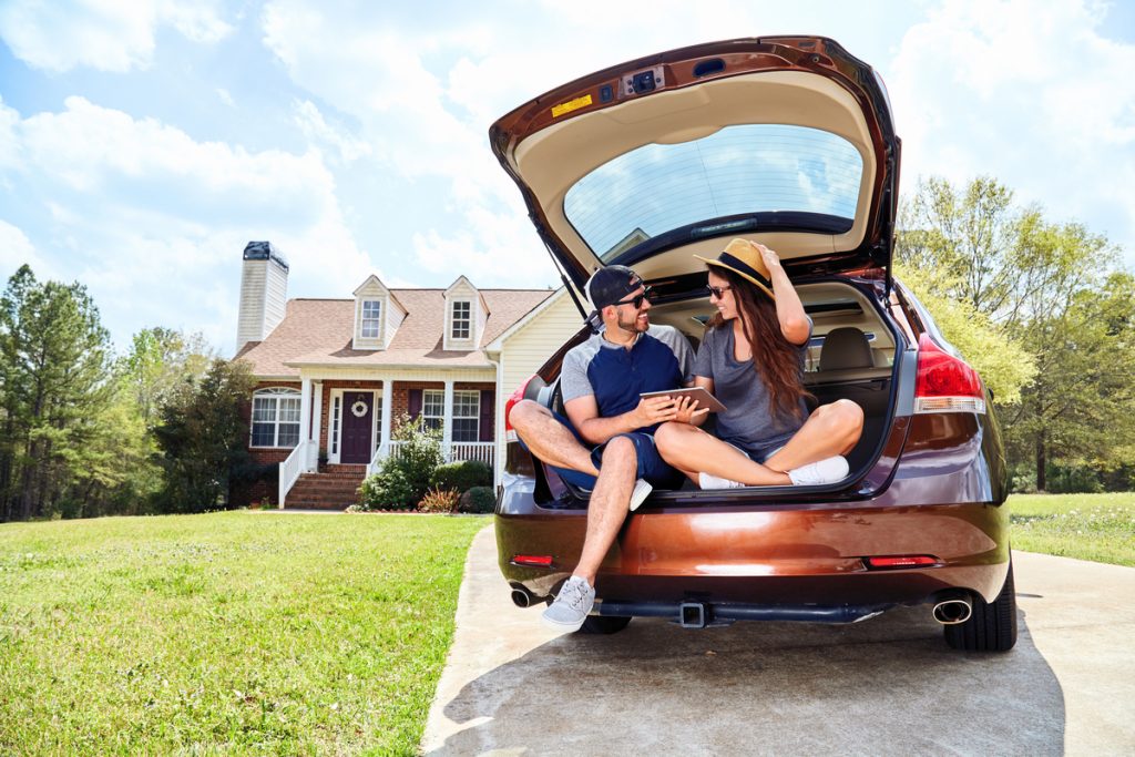 Man and woman sitting in the back of their car in the driveway of their house.