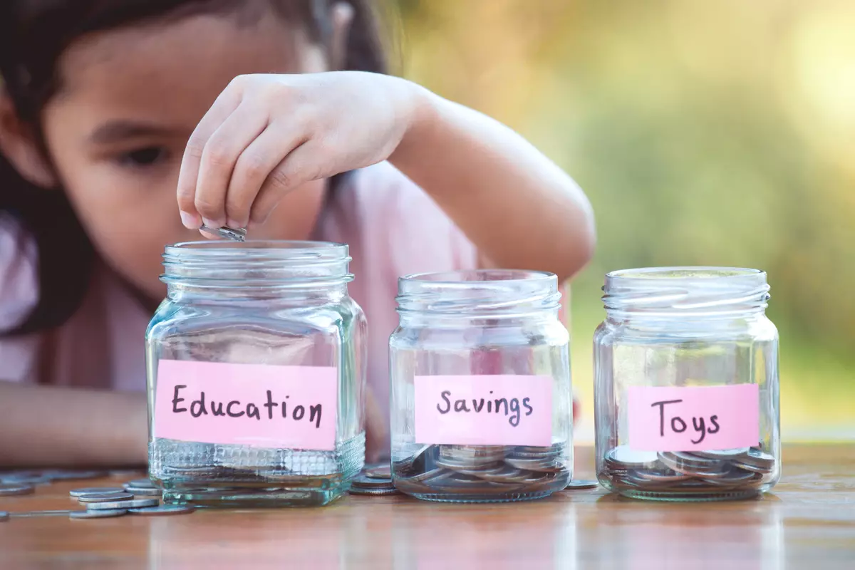 A child dropping coins into three different jars, labelled "education," "savings," and "toys."