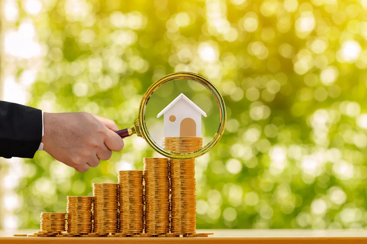 A hand holding a magnifying glass over a house to investigate property value.