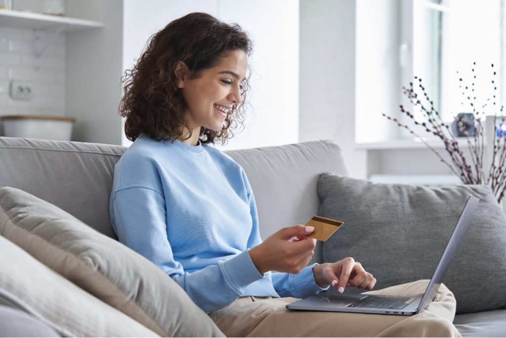 Smiling young adult sitting on couch with laptop and holding credit card.