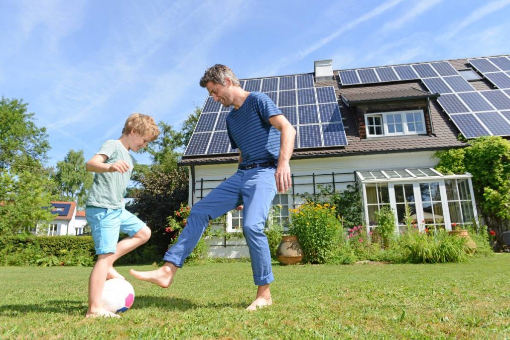 Father and son playing soccer in front house with solar panels.